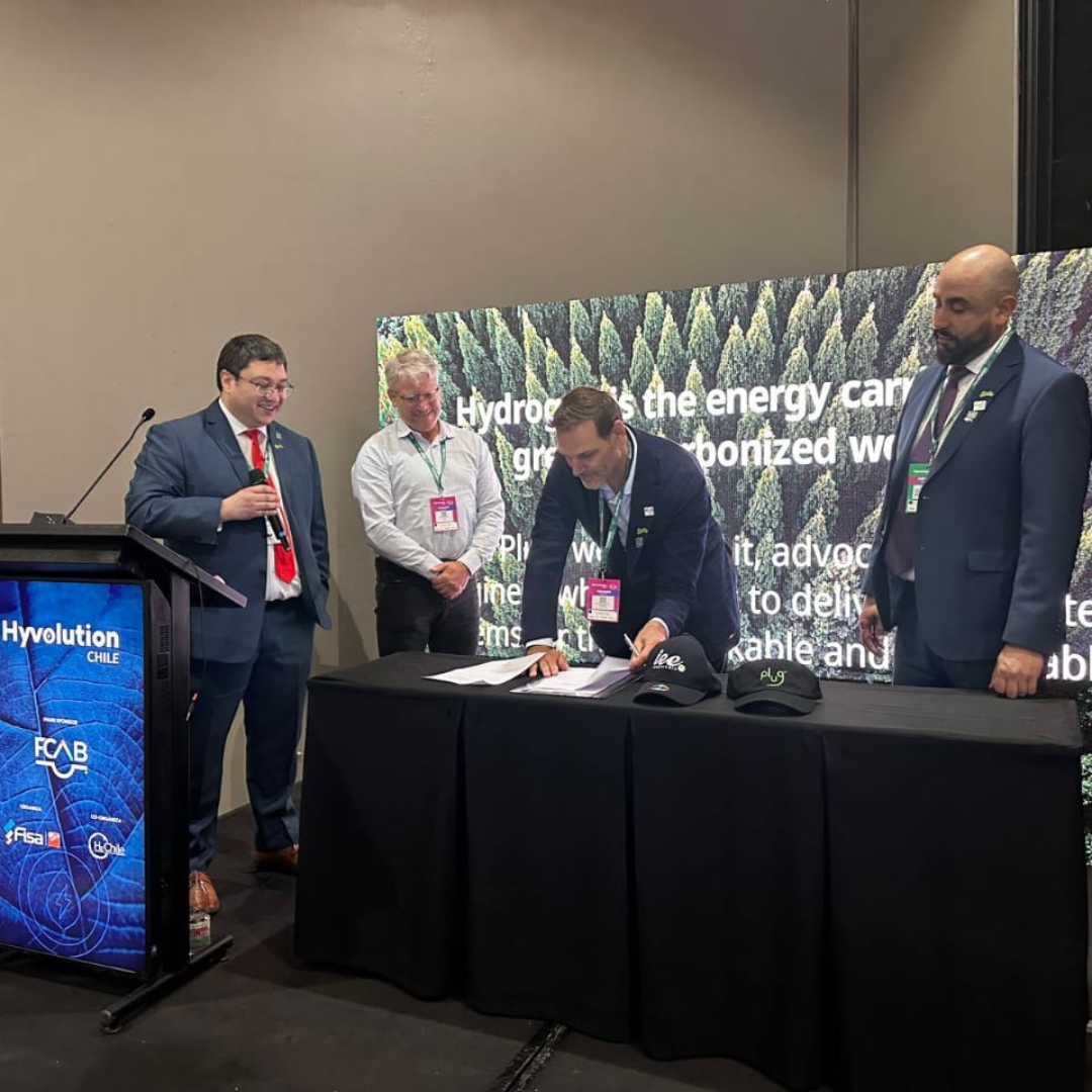 Quatro executivos participam de uma cerimônia de assinatura de parceria na Hyvolution Chile. Um deles está assinando o documento em uma mesa coberta por toalha preta, enquanto os outros observam. Ao fundo, um grande painel com imagem de floresta e frases sobre hidrogênio verde destaca o tema de sustentabilidade e transição energética.