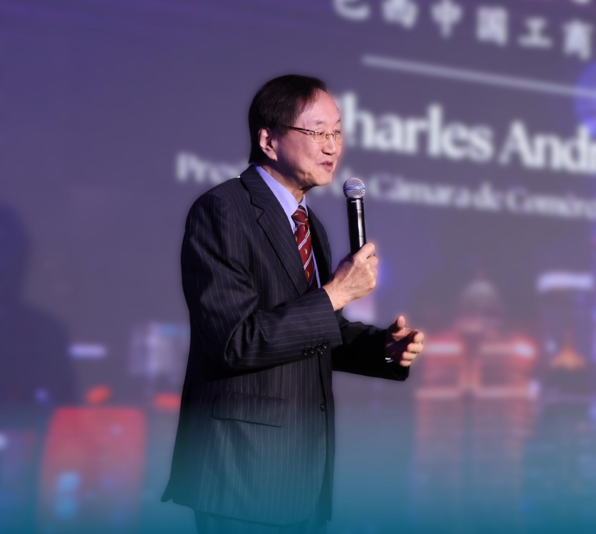 Charles Tang em evento do Tang International Symposium 2025 segurando um microfone e falando no palco, com fundo projetado relacionado ao setor de comércio e desenvolvimento sustentável.